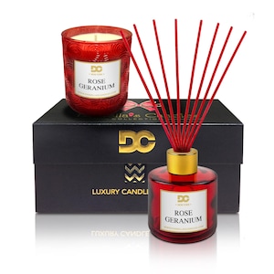 May include: A black gift box with a gold logo and the words "Luxury Candles" on the front. The box contains a red glass candle and a red glass diffuser with gold accents. Both the candle and diffuser have the words "Rose Geranium" printed on the front.
