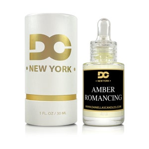 May include: A white and gold cylindrical box and a clear glass bottle of Amber Romancing oil. The box has the text "DC NEW YORK" in gold. The bottle has a black label with the text "AMBER ROMANCING" and "1 FL. OZ / 30 ML".