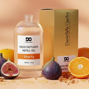 May include: A clear glass bottle of Citrus Fig reed diffuser refill oil with a white label reading DC New York, 100mL 3.4oz, next to a gold box labeled Daniella's Candles Collection, surrounded by fresh whole and halved figs and orange slices.