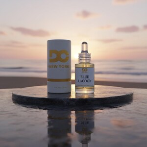 Puede incluir: Una botella de vidrio transparente de aceite "Blue Lagoon" con un cuentagotas, junto a una caja cil&iacute;ndrica blanca con letras doradas. La caja dice "DC NEW YORK" y "1 fl oz / 30 ml". El fondo es una playa al atardecer.