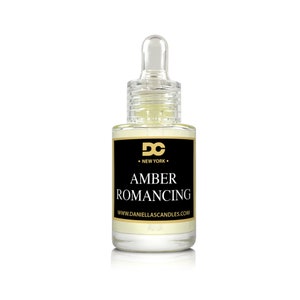 May include: A clear glass bottle with a dropper, filled with a light amber liquid. The bottle has a black label with gold lettering that reads "AMBER ROMANCING" and "DC NEW YORK". The website address www.daniellascandles.com is also visible.