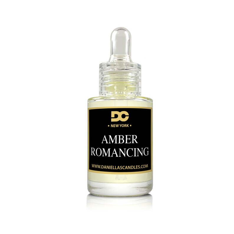 May include: A clear glass bottle with a dropper, filled with a light amber liquid. The bottle has a black label with gold lettering that reads "AMBER ROMANCING" and "DC NEW YORK". The website address www.daniellascandles.com is also visible.