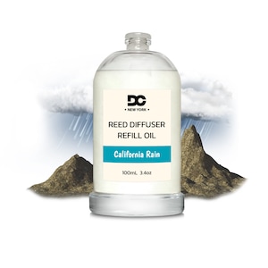 May include: A clear glass bottle filled with a light-colored liquid, labeled "California Rain" reed diffuser refill oil. The label includes the text "DC New York" and the bottle contains 3.4 oz of oil. The background features a rain cloud and two brown rock formations.