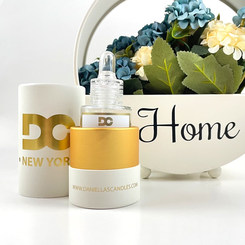 Puede incluir: Una botella de vidrio transparente con una etiqueta dorada y blanca y una tapa dorada. La botella est&aacute; junto a un cilindro blanco con letras doradas que dice "DC NEW YORK". Un macetero blanco con la palabra "Home" y flores est&aacute; en el fondo.