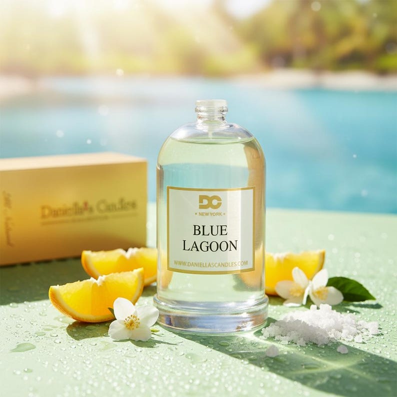 Puede incluir: Una vela de cristal transparente con una etiqueta dorada que dice "Blue Lagoon" y "DC New York". La vela est&aacute; rodeada de rodajas de naranja, flores blancas y sal marina. Una caja dorada est&aacute; en el fondo.