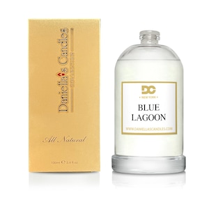 Puede incluir: Una vela de cristal transparente con una etiqueta blanca que dice "Blue Lagoon". La vela est&aacute; junto a una caja dorada con el texto "Daniella's Candles Collection" y "All Natural". La vela contiene 100 ml.