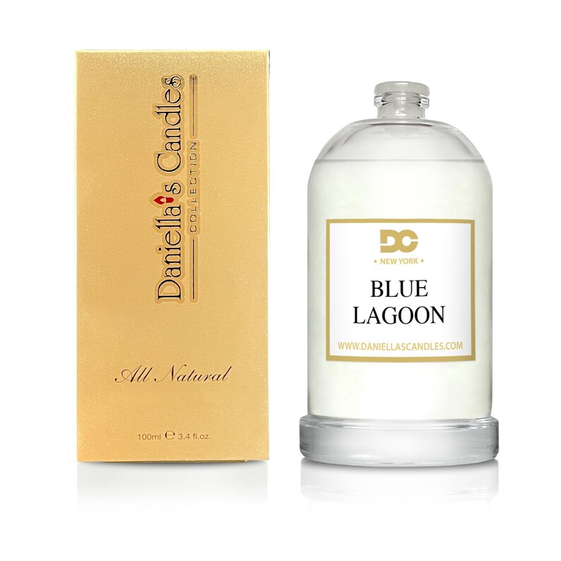 Puede incluir: Una vela de cristal transparente con una etiqueta blanca que dice "Blue Lagoon". La vela est&aacute; junto a una caja dorada con el texto "Daniella's Candles Collection" y "All Natural". La vela contiene 100 ml.