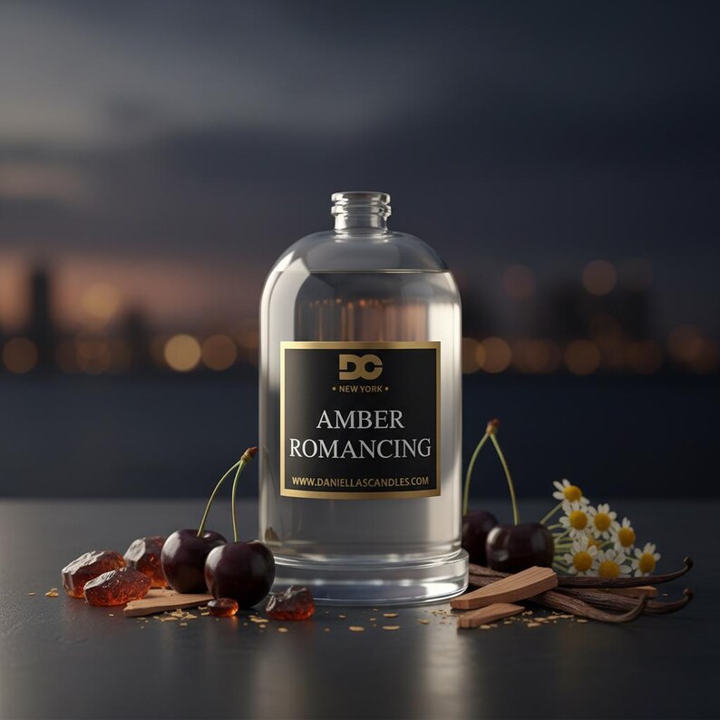 May include: A clear glass bottle with a black and gold label that reads "Amber Romancing." The bottle is surrounded by cherries, amber-colored crystals, vanilla beans, and small white flowers. The background is blurred.