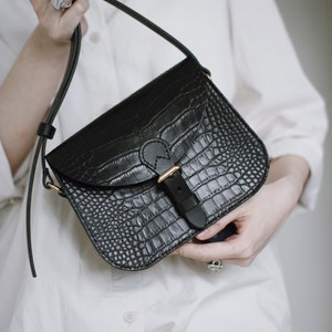 May include: A black leather crossbody bag with a crocodile-embossed texture and a gold buckle closure.