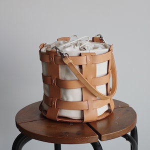Pode incluir: Uma bolsa de couro marrom em forma de balde com forro de lona branca e uma alça de couro trançada. A bolsa está em cima de um banquinho de madeira.