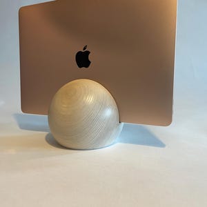 May include: A rose gold laptop with a black Apple logo is held by a light-colored wooden spherical stand. The stand has a natural wood grain pattern. The laptop is angled slightly, showcasing its sleek design.
