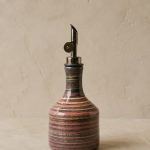 Op de afbeelding: Een keramische oliefles met een glanzende afwerking en horizontale strepen in tinten donkerbruin, gedempt rood en crème. De fles is voorzien van een zilverkleurige metalen schenktuit met een scharnierend deksel voor gecontroleerd schenken.