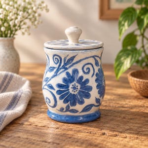 Op de afbeelding: Een keramische voorraadpot met deksel, voorzien van een witte basis versierd met handgeschilderde blauwe bloemmotieven en krullende ranken. De cilindrische pot heeft een uitlopende voet, een blauwe rand en een knop op het deksel.