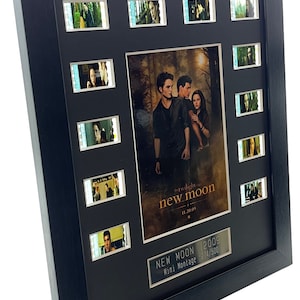 May include: A black framed movie cell montage of the Twilight Saga: New Moon. The montage features 10 film cells with scenes from the movie. The poster for the movie is in the center of the frame. The text on the poster reads "The Twilight Saga: New Moon" and "11.20.09". The text on the plaque below the poster reads "New Moon [2009] Mini Montage 274/500".