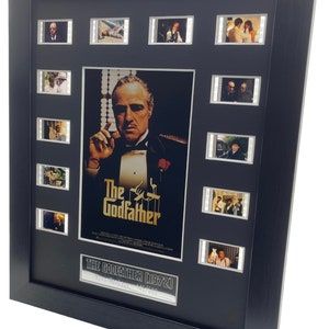Puede incluir: Un p&oacute;ster de pel&iacute;cula enmarcado de El Padrino con un marco negro y una placa plateada que dice "The Godfather 1972, Film Montage 146/500". El p&oacute;ster presenta un primer plano de Marlon Brando como Don Vito Corleone. Hay 10 tiras de celdas de pel&iacute;cula alrededor del p&oacute;ster.