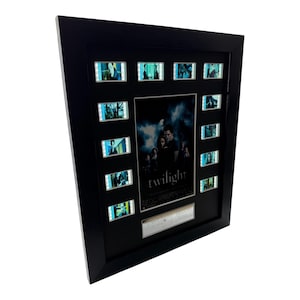 May include: A black framed display featuring a central Twilight movie poster surrounded by twelve individual film cells. The poster shows characters in a dark, moody setting with the word twilight printed in white text at the bottom centre.