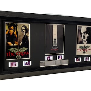 Könnte beinhalten: Eine schwarze gerahmte Film-Memorabilia-Anzeige mit drei Filmplakaten von The Crow und Filmzellenstreifen. Die Poster zeigen ein Schwarzweißbild von Brandon Lee, ein Ganzkörperbild von Brandon Lee und eine Nahaufnahme von Brandon Lees Gesicht. Die Filmzellenstreifen sind in Schwarzweiß.