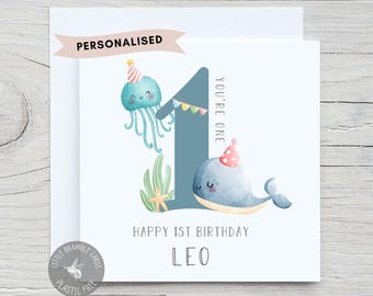 May include: A square greeting card featuring a large blue number one, a teal jellyfish, and a blue whale, both wearing party hats. The card includes the text "YOU'RE ONE", "HAPPY 1ST BIRTHDAY", and the name "LEO" in a simple sans-serif font.