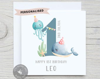 Op de afbeelding: Gepersonaliseerde kaart voor de 1e verjaardag met een blauwe walvis en een kwal die feestmutsen dragen. De kaart staat "You're one" en "Happy 1st Birthday Leo".
