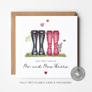 Pode incluir: Um cartão branco com uma ilustração em aquarela de dois pares de botas de chuva, uma preta e outra rosa com bolinhas brancas. O texto "Nosso primeiro ano como Sr. e Sra. Watts" está impresso abaixo das botas. O cartão é rodeado por um envelope marrom.