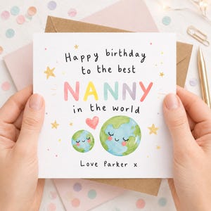 K&ouml;nnte beinhalten: Eine quadratische Gru&szlig;karte mit wei&szlig;em Hintergrund und dem Text Happy birthday to the best NANNY in the world in schwarzen und bunten Buchstaben. Unter dem Text befinden sich zwei l&auml;chelnde Erden, ein rosa Herz und goldene Sterne.
