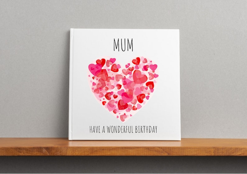 May include: A white card with the word "MUM" in black text and a large pink and red heart made of smaller hearts. The text "HAVE A WONDERFUL BIRTHDAY" is below the heart.