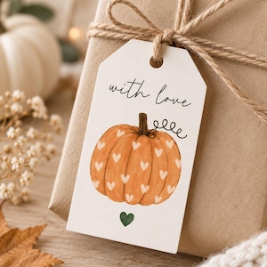 Op de afbeelding: Een wit rechthoekig cadeaulabel vastgebonden met bruin touw, met de tekst "with love" in zwarte schrijfletters boven een illustratie van een oranje pompoen versierd met kleine witte hartjes en één klein groen hartje onderaan.