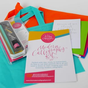 May include: A modern calligraphy kit in a turquoise tote bag. The kit includes a calligraphy pen, ink, and a booklet with instructions. The booklet has the title "Modern Calligraphy Kit" in pink script. The tote bag is next to a green tote bag with a white card that says "Because of you, I laugh a little louder."