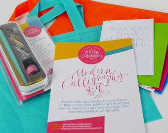 May include: A modern calligraphy kit in a turquoise tote bag. The kit includes a calligraphy pen, ink, and a booklet with instructions. The booklet has the title "Modern Calligraphy Kit" in pink script. The tote bag is next to a green tote bag with a white card that says "Because of you, I laugh a little louder."