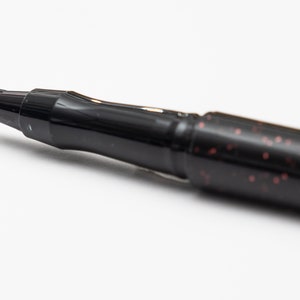 May include: Close-up of a black brush pen with a fine tip. The pen has a black barrel with a subtle red glitter finish.
