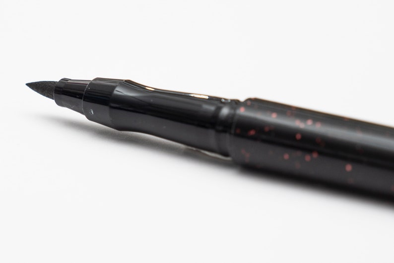 May include: Close-up of a black brush pen with a fine tip. The pen has a black barrel with a subtle red glitter finish.