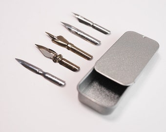 May include: A set of five silver and gold calligraphy nibs in a silver metal tin. The nibs are arranged in a row on a white surface.