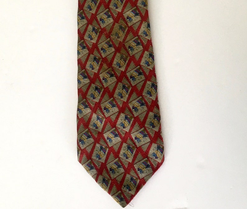 May include: A patterned necktie featuring a repeating geometric design. The tie has a red background with a diamond pattern in a lighter shade of red. Within each diamond are smaller squares with a floral pattern in shades of blue, yellow, and purple.