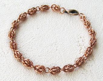 May include: A copper bracelet with a woven chain design. The bracelet is made of small, interconnected rings that create a delicate and intricate pattern.
