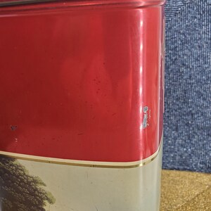 May include: A vintage red metal tin with a lid. The tin features a landscape scene with a horse-drawn carriage and figures. The top and lid are a solid red colour, with a gold trim separating the red from the landscape scene.