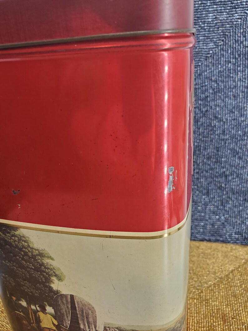 May include: A vintage red metal tin with a lid. The tin features a landscape scene with a horse-drawn carriage and figures. The top and lid are a solid red colour, with a gold trim separating the red from the landscape scene.