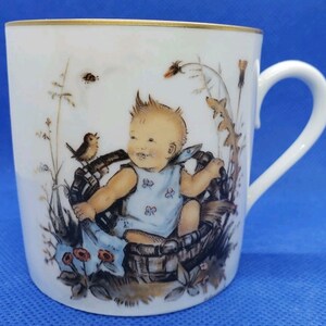 Può includere: Una tazza in porcellana bianca con un bordo dorato presenta un'illustrazione colorata di un bambino seduto in un cesto con un uccello appollaiato sul bordo. Il bambino indossa un vestito blu e bianco con un motivo floreale. Lo sfondo è un paesaggio verde e marrone con fiori e foglie.