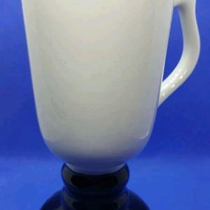 May include: A white ceramic mug with a black base and stem. The mug has a curved handle and a wide opening. The mug is set against a blue background.