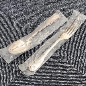 May include: Two pieces of Christofle silverware, a spoon and a fork, in their original packaging. The silver utensils are sealed in clear plastic. The spoon and fork are on a textured, dark blue surface.