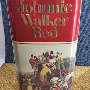 May include: A red Johnnie Walker Red tin with gold lettering. The lower section displays a vintage illustration of a horse-drawn carriage and figures in historical attire. The tin is rectangular and tall.