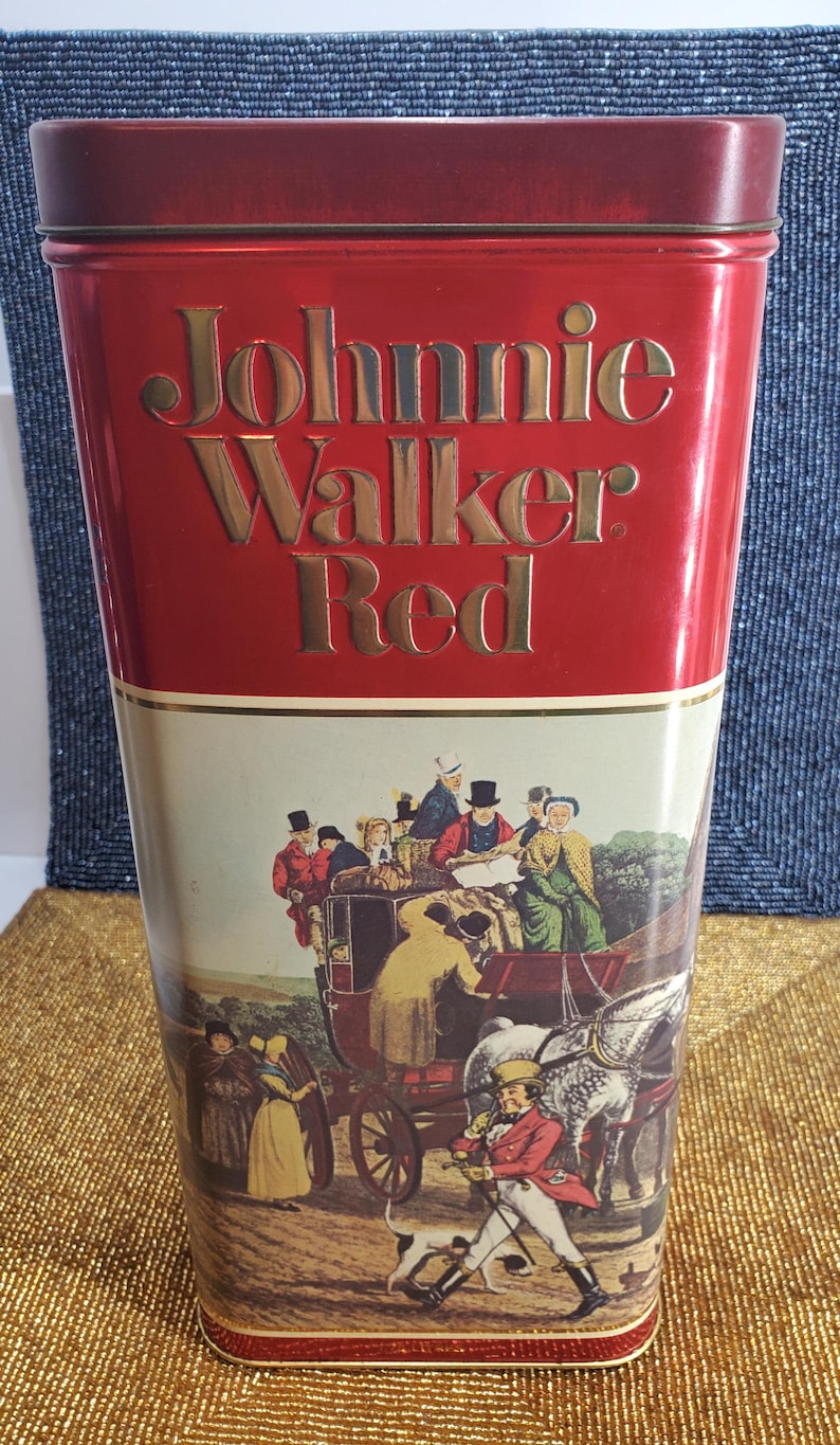 May include: A red Johnnie Walker Red tin with gold lettering. The lower section displays a vintage illustration of a horse-drawn carriage and figures in historical attire. The tin is rectangular and tall.