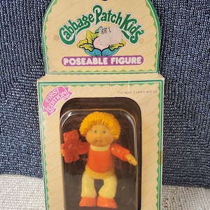 Könnte beinhalten: Eine bewegliche Cabbage Patch Kids Figur in der Originalverpackung. Die Puppe mit gelben Haaren, Sparky Patrick, trägt ein orange-gelbes Outfit und hält einen kleinen braunen Bären. Die Schachtel zeigt den Text First Edition und Hello, My Name Is Sparky Patrick.