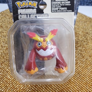 May include: A Pokemon Figure Collection toy, featuring a red and orange character with yellow accents, packaged in a clear plastic case. The packaging includes the Pokemon logo and a warning about choking hazards and sharp points.