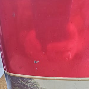May include: A vintage red metal container with a landscape design at the bottom. The container has a rectangular shape and a lid. The red paint shows signs of wear, adding to its antique charm. The landscape features trees and a field.