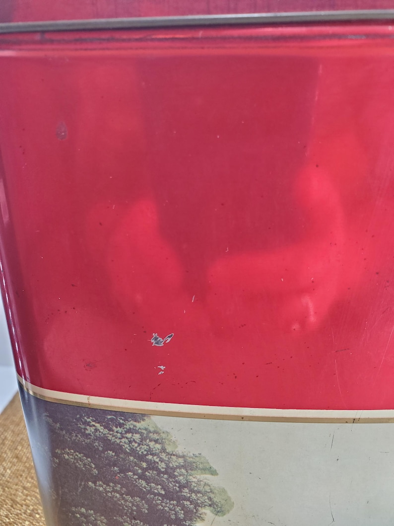May include: A vintage red metal container with a landscape design at the bottom. The container has a rectangular shape and a lid. The red paint shows signs of wear, adding to its antique charm. The landscape features trees and a field.