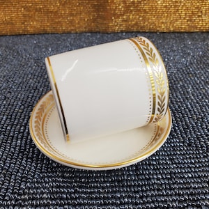 Puede incluir: Juego de taza y platillo blancos con hojas doradas y ribete punteado. La taza está inclinada, revelando el interior. El platillo tiene un diseño de hojas doradas a juego alrededor del borde. El juego está sobre una superficie de cuentas azul oscuro.