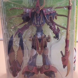 Może przedstawiać: Czerwono-złota figurka akcji wojownika z mieczem i laską. Figurka znajduje się w plastikowym opakowaniu z napisem "Ultra-Action Figures" z przodu.