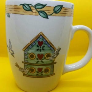 May include: White ceramic mug with a birdhouse design. The birdhouse is green with red heart accents and birds perched on it. A band of brown and yellow with green leaves decorates the top of the mug. The mug has a white handle.