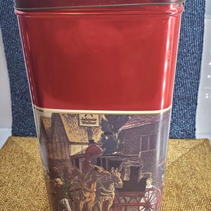 May include: A red metal tin with a rectangular shape and a hinged lid. The lower section features a vintage illustration of a horse-drawn carriage with the "Johnnie Walker" logo.