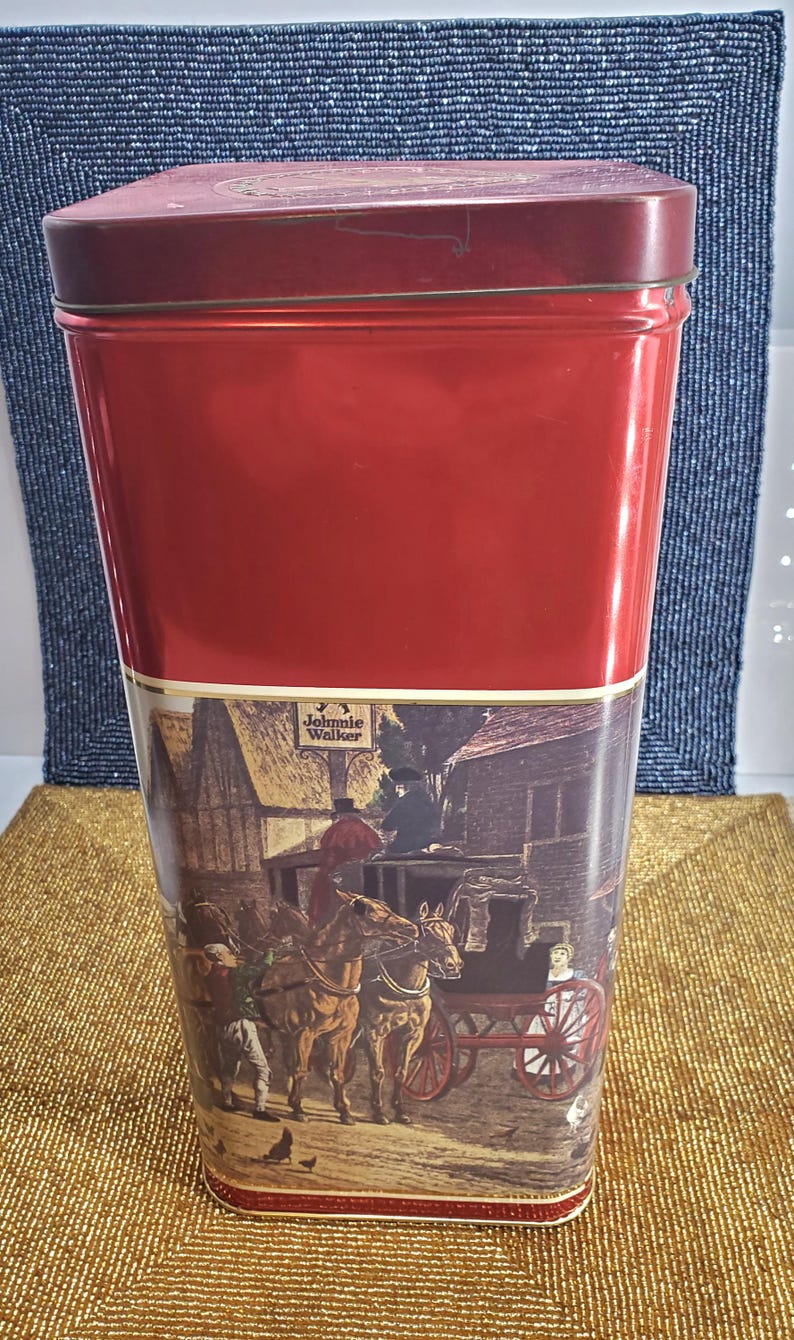 May include: A red metal tin with a rectangular shape and a hinged lid. The lower section features a vintage illustration of a horse-drawn carriage with the "Johnnie Walker" logo.